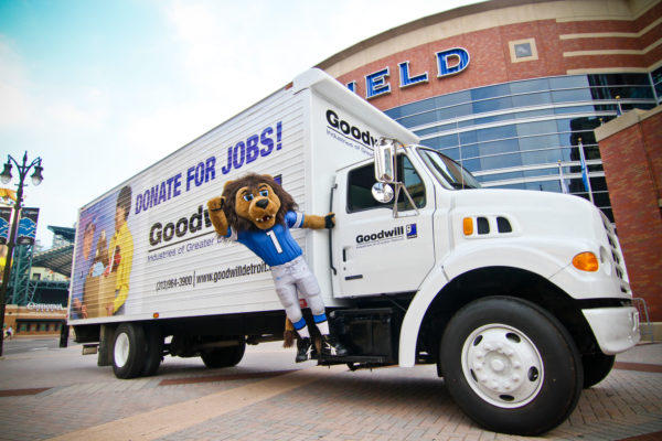 Image of Goodwill truck with Detroit lion mascot hanging off it in front of Ford field