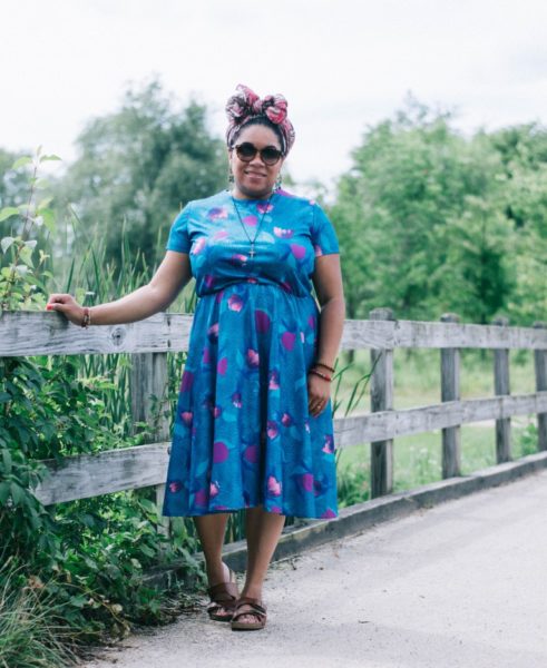 Woman wearing sunglasses, blue vintage dress, and colorful head wrap leaning on a fence.